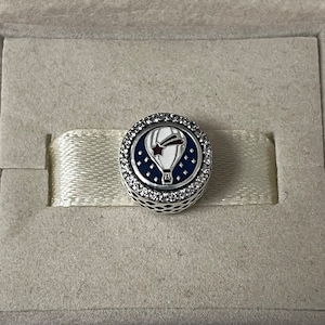 May include: A silver charm with a white hot air balloon with a red star and a blue comet tail on a blue background with white stars. The charm is surrounded by a circle of clear cubic zirconia stones.
