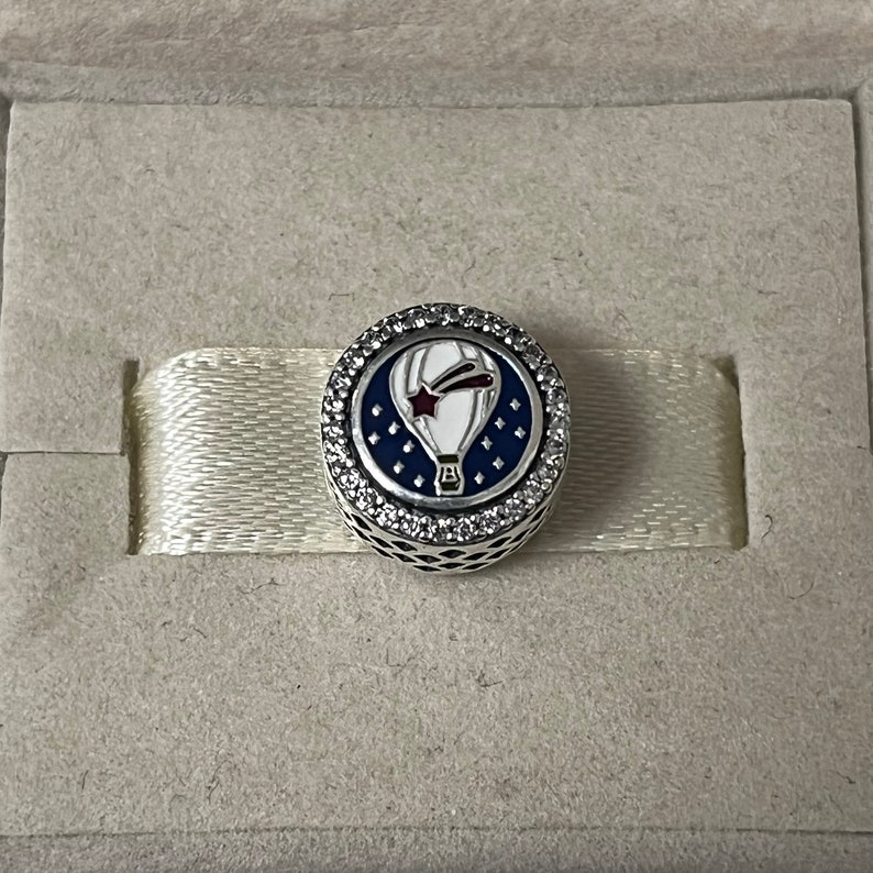 May include: A silver charm with a white hot air balloon with a red star and a blue comet tail on a blue background with white stars. The charm is surrounded by a circle of clear cubic zirconia stones.