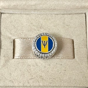 May include: A silver charm with a blue and yellow enamel design of the flag of Barbados. The charm is surrounded by a circle of clear cubic zirconia stones.