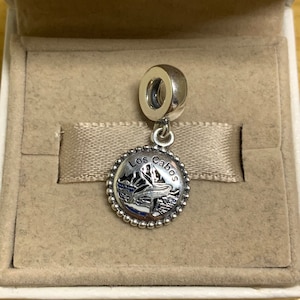 May include: A silver charm with a blue and white enamel design of a mountain range and the text "Los Cabos".