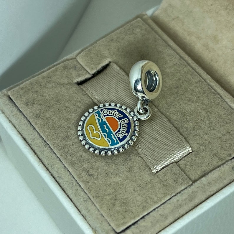 K&ouml;nnte beinhalten: Silberner Anh&auml;nger mit einem blauen, gelben und orangefarbenen Emaille-Design, das einen Sonnenuntergang &uuml;ber dem Meer darstellt. Der Anh&auml;nger tr&auml;gt die Worte "Outer Banks" um den Rand herum.