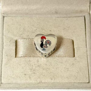 May include: A heart-shaped silver charm with a painted rooster design. The rooster has a red comb, black body, and a dotted pattern on its tail. The charm is presented in a beige jewelry box with a satin ribbon.