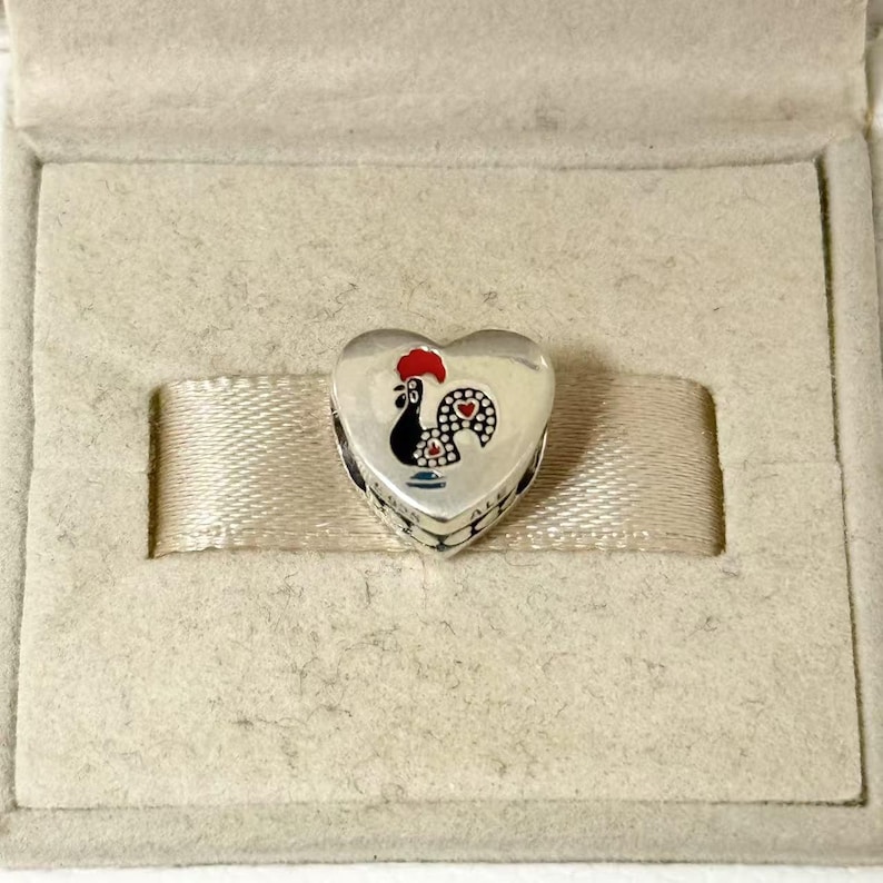May include: A heart-shaped silver charm with a painted rooster design. The rooster has a red comb, black body, and a dotted pattern on its tail. The charm is presented in a beige jewelry box with a satin ribbon.