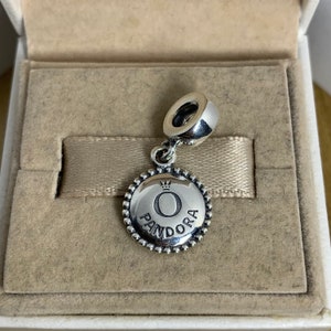 May include: Silver charm with a round, textured disc featuring the Pandora logo and a crown above the letter "O".