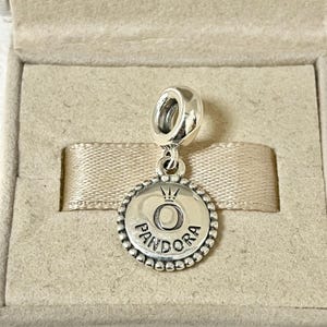 Pu&ograve; includere: Un ciondolo d'argento con un bordo rotondo e perlato. Il ciondolo presenta una corona sopra la lettera "O" e la parola "PANDORA" sotto.