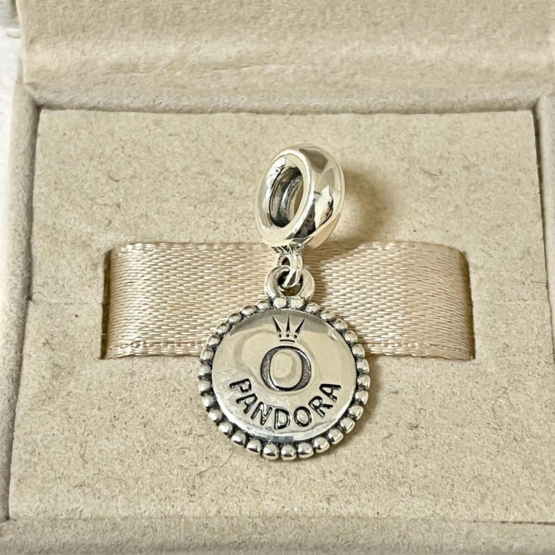 Pu&ograve; includere: Un ciondolo d'argento con un bordo rotondo e perlato. Il ciondolo presenta una corona sopra la lettera "O" e la parola "PANDORA" sotto.