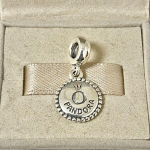 May include: Silver charm with a round, beaded border. The charm features a raised, silver letter "O" with a crown above it. The word "PANDORA" is inscribed below the letter.