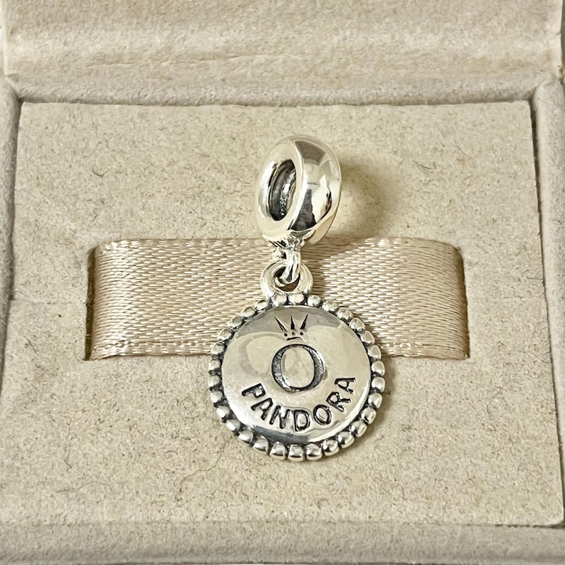 May include: Silver charm with a round, beaded border. The charm features a raised, silver letter "O" with a crown above it. The word "PANDORA" is inscribed below the letter.