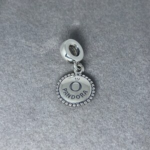 May include: Silver charm with a round, textured disc featuring the Pandora logo, a crown, and the letter "O".