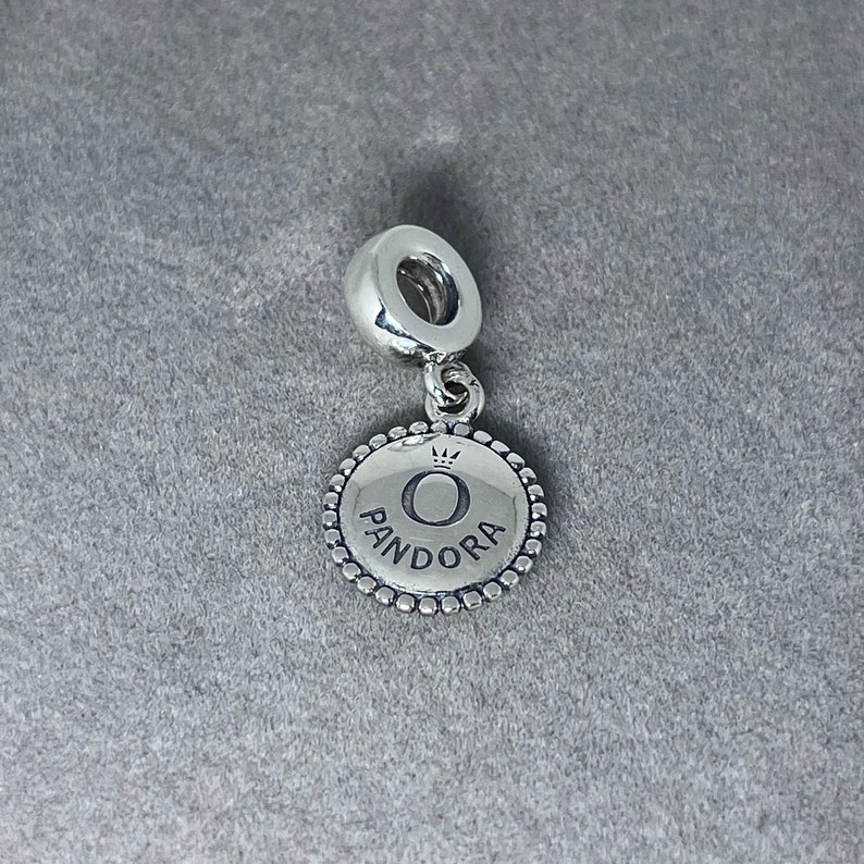 May include: Silver charm with a round, textured disc featuring the Pandora logo, a crown, and the letter "O".