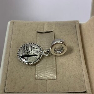 Puede incluir: Encanto de plata con un dise&ntilde;o circular que presenta una imagen en blanco y negro de un edificio con una bandera estadounidense. El encanto tiene un peque&ntilde;o bucle para sujetarlo a una pulsera.