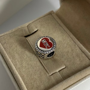 May include: A silver charm in the shape of a red heart with the letters NYC in white on it. The heart is surrounded by a circle of clear cubic zirconia stones.