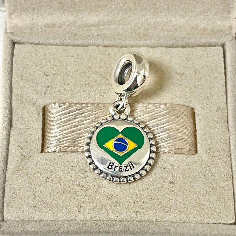 May include: Silver charm with a green, yellow, and blue heart-shaped enamel design representing the Brazilian flag. The charm has a beaded border and the word "Brazil" inscribed below the heart.