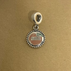 May include: Silver charm with a peach design and the word "GEORGIA" in black lettering. The peach is pink and has a green leaf.