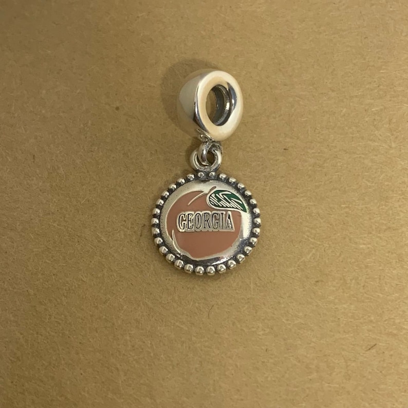 May include: Silver charm with a peach design and the word "GEORGIA" in black lettering. The peach is pink and has a green leaf.