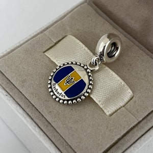 Op de afbeelding: Zilveren bedel met een rond blauw en geel emaille ontwerp van de vlag van Barbados. De bedel heeft een zilveren kralenrand en het woord "Barbados" staat onderaan.
