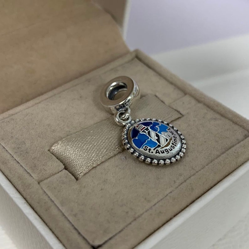 K&ouml;nnte beinhalten: Ein silberner Anh&auml;nger mit einem blauen und wei&szlig;en Emaille-Design eines Leuchtturms und dem Text "St. Augustine" in einem wei&szlig;en Kreis mit einem silbernen Perlenrand.
