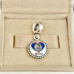 May include: A circular silver charm featuring a heart-shaped enamel inlay with the blue, white, and red colours of the Belize flag. The centre displays the national coat of arms, and the word Belize is printed in black below the heart design.