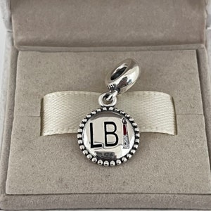 May include: Silver charm with a round, beaded border. The charm features a white circle with the letters "LB" in black and a red and white design on the right side.