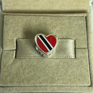 May include: A silver heart charm with a red, black, and white enamel design representing the flag of Trinidad and Tobago.