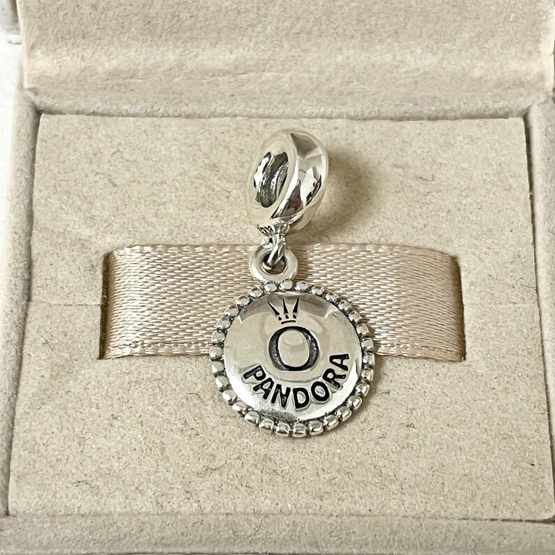 May include: A silver charm with a round, silver charm with a black 'O' and the word 'PANDORA' around it. The charm has a small crown above the 'O'.