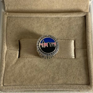 Puede incluir: Un charm de plata con un dise&ntilde;o de ciudad en blanco y negro. El charm est&aacute; rodeado por un c&iacute;rculo de piedras de circonita c&uacute;bica transparentes. La ciudad est&aacute; sobre un fondo rosa, azul y negro.