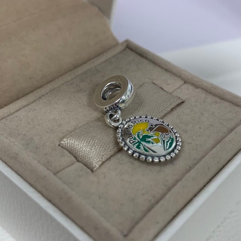 May include: A silver charm with a round pendant featuring a colourful design of Puerto Rico. The charm has a loop at the top for attaching to a bracelet or necklace. The pendant has a beaded edge and the words "Puerto Rico" around the edge.