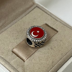 May include: A silver charm featuring a red and white enamel Turkish flag design. The charm is encircled by a ring of clear cubic zirconia stones.
