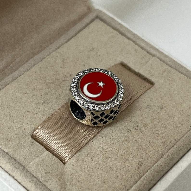 May include: A silver charm featuring a red and white enamel Turkish flag design. The charm is encircled by a ring of clear cubic zirconia stones.