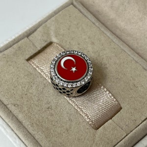 May include: A silver charm featuring a red enamel Turkish flag with a white crescent moon and star. The charm is surrounded by a border of clear cubic zirconia stones.