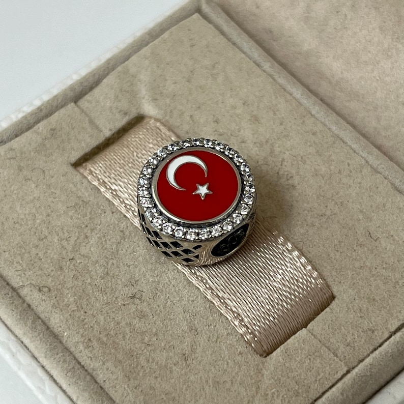May include: A silver charm featuring a red enamel Turkish flag with a white crescent moon and star. The charm is surrounded by a border of clear cubic zirconia stones.