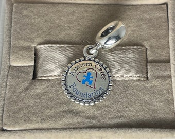 May include: Silver charm with a blue and red heart with a puzzle piece inside. The charm says "Autism Cares Foundation" around the heart.
