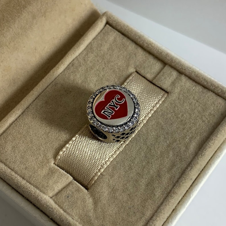 May include: A silver charm featuring a red heart with the letters NYC in black. The charm is encircled by a ring of clear cubic zirconia stones.
