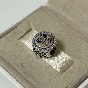 May include: A silver charm with a clear crystal border, featuring a colorful illustration of a couple in traditional clothing. The charm is in a white box with a textured surface.