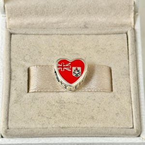 May include: A silver heart-shaped charm with a red enamel design of the Bermuda flag. The flag features a red field with a British Union Jack in the upper hoist quadrant and the Bermuda coat of arms in the lower fly quadrant.