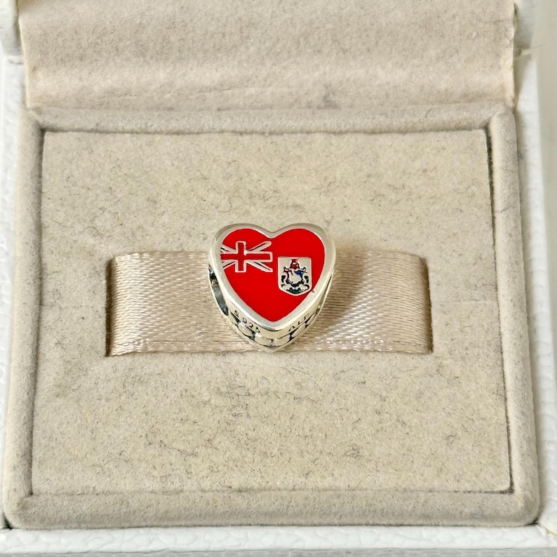 May include: A silver heart-shaped charm with a red enamel design of the Bermuda flag. The flag features a red field with a British Union Jack in the upper hoist quadrant and the Bermuda coat of arms in the lower fly quadrant.