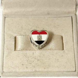May include: Heart-shaped charm with the colors of the Egyptian flag: red, white, and black. The charm has a silver border and features an emblem in the center. The charm is inside a beige box.