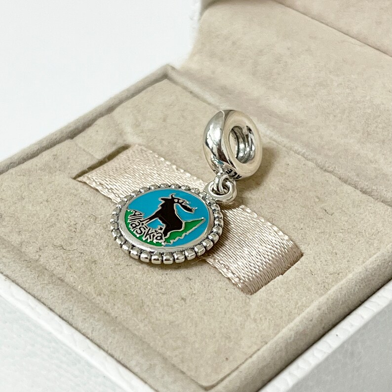May include: A silver charm with a blue and green enamel design of a mountain goat and the word "Alaska" in a circle. The charm is in a white box.