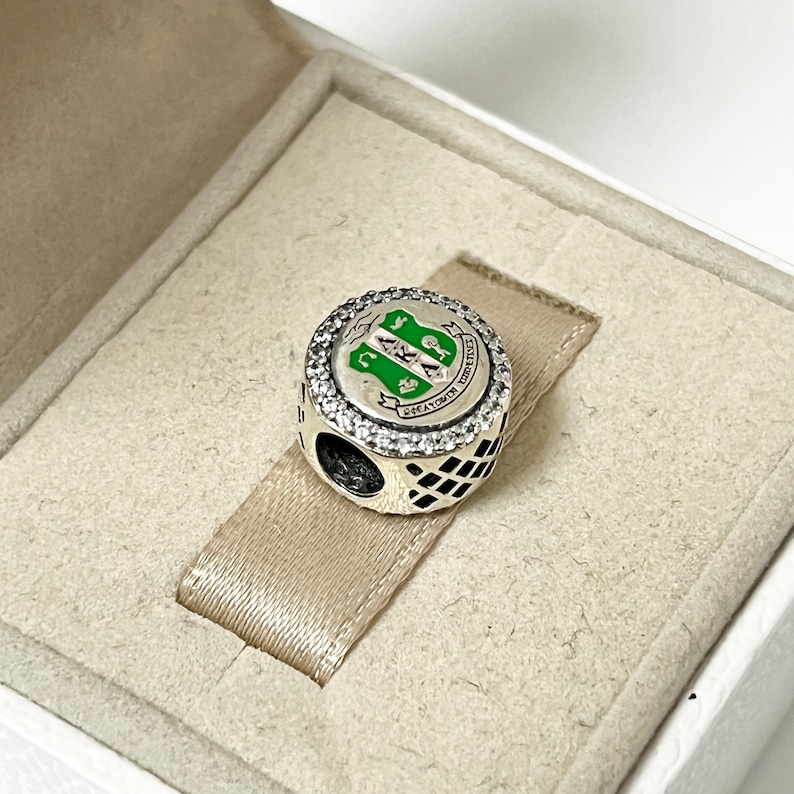 May include: A silver charm with a green and white enamel design featuring the letters "AKA" and the words "AKA Sorority, Inc." surrounded by clear cubic zirconia stones.