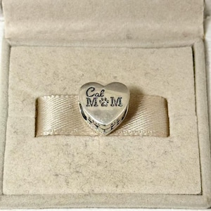 Puede incluir: Un charm de plata en forma de coraz&oacute;n con la inscripci&oacute;n "Cal M&M" y huellas de patas. El charm se presenta en una caja de joyer&iacute;a beige con una cinta de sat&eacute;n.