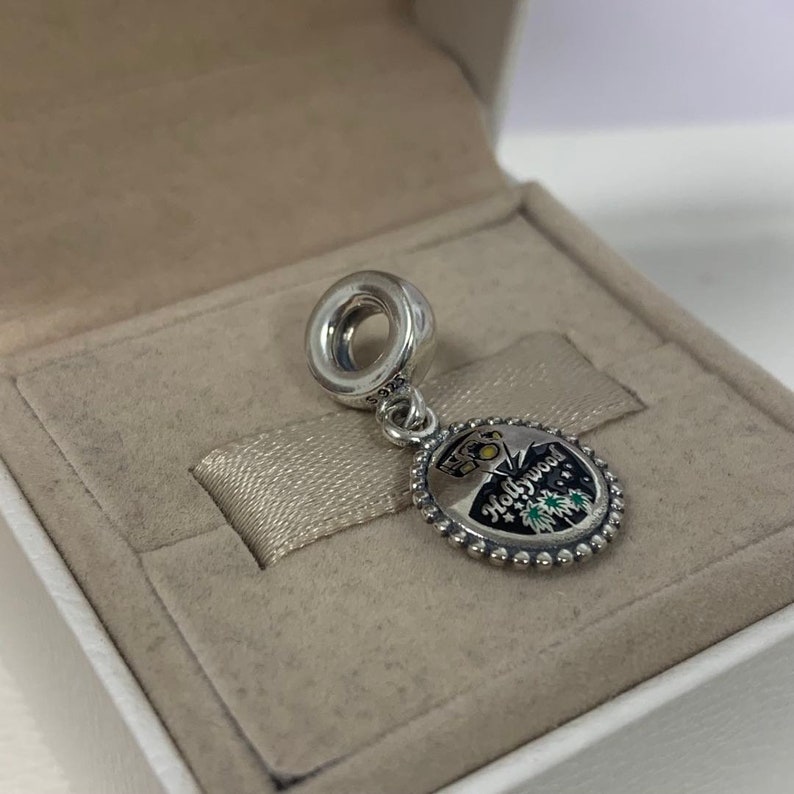 May include: Silver charm featuring a black and white enamel design of the Hollywood sign and palm trees. The charm has a silver ring at the top for attaching to a bracelet.