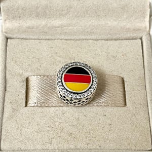 Könnte beinhalten: Ein silberner Anhänger mit einem schwarzen, roten und gelben Emaille-Design der deutschen Flagge. Der Anhänger ist von einem Kreis aus klaren Zirkonia-Steinen umgeben.