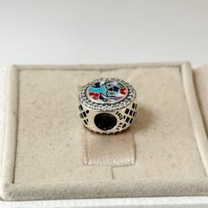 May include: A silver charm with a colorful enamel design of Stitch from Lilo & Stitch. The charm is surrounded by clear cubic zirconia stones and has the text "925 ALE" on the back.