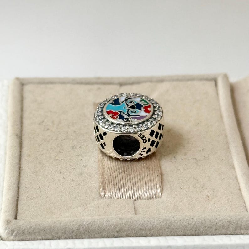 May include: A silver charm with a colorful enamel design of Stitch from Lilo & Stitch. The charm is surrounded by clear cubic zirconia stones and has the text "925 ALE" on the back.