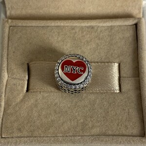 May include: A silver charm with a red heart featuring the text "NYC" surrounded by clear cubic zirconia stones.