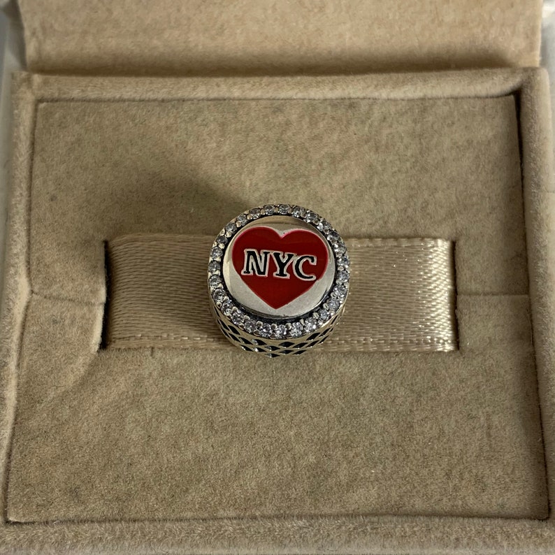 May include: A silver charm with a red heart featuring the text "NYC" surrounded by clear cubic zirconia stones.