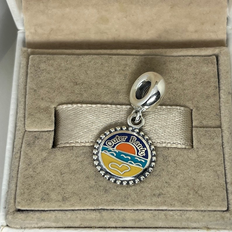 K&ouml;nnte beinhalten: Silberner Anh&auml;nger mit einem blauen, gelben und orangefarbenen Emaille-Design einer Strandszene mit einem Herzen und den Worten "Outer Banks".