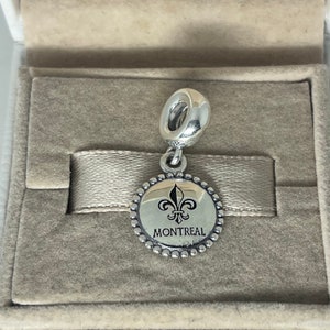 May include: Silver charm with a round, silver-colored disc featuring a black fleur-de-lis and the word "MONTREAL" in black lettering. The charm has a silver loop for attaching to a bracelet.