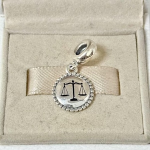 May include: A silver charm with a round pendant featuring a black scale of justice symbol. The charm has a beaded border and a loop for attaching to a bracelet or necklace. The charm is displayed in a beige jewelry box.