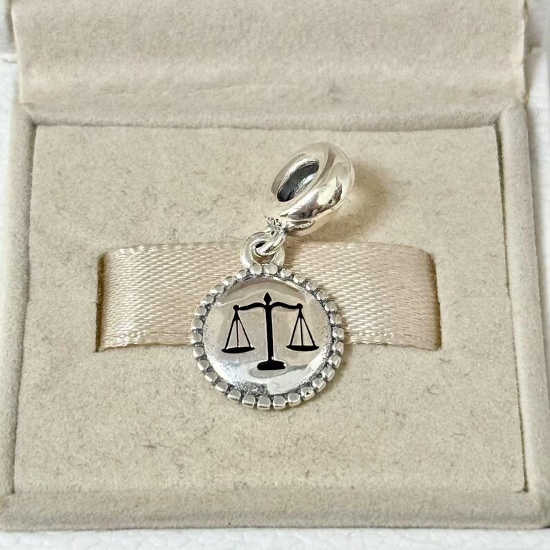 May include: A silver charm with a round pendant featuring a black scale of justice symbol. The charm has a beaded border and a loop for attaching to a bracelet or necklace. The charm is displayed in a beige jewelry box.